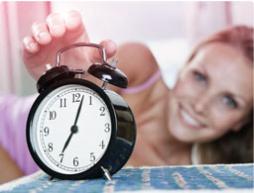 Billede af et vækkeur med en kvinde i baggrunden, der strækker sig for at slukke alarmen.